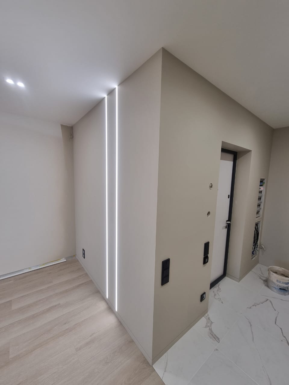 Nowoczesne wnętrze przedpokoju z dwiema pionowymi liniami oświetlenia LED w ścianie, jasnymi panelami podłogowymi i marmurową posadzką przy drzwiach wejściowych. Widoczna puszka farby w rogu.
