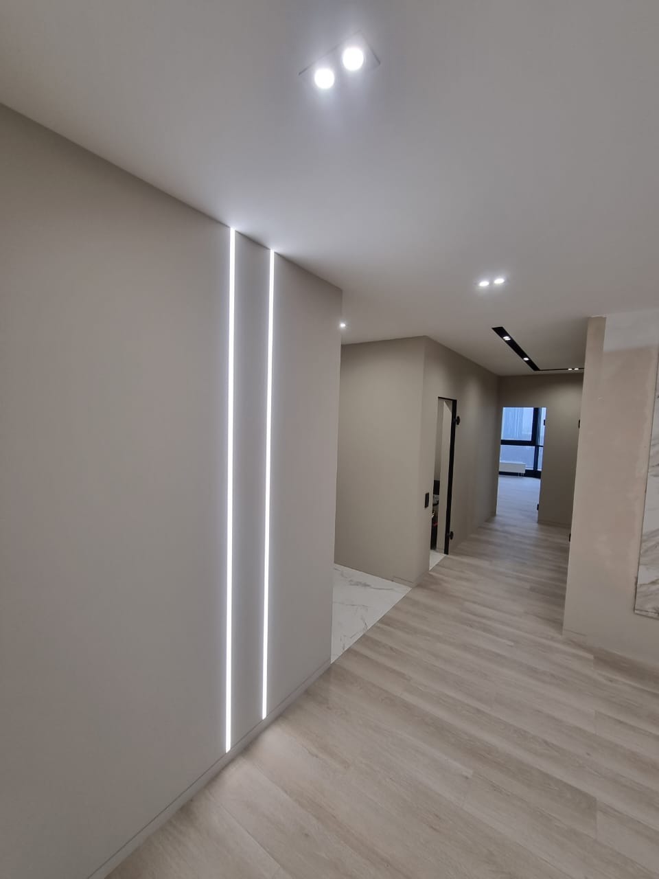 Nowoczesny korytarz z dwiema pionowymi liniami LED w ścianie, jasne panele podłogowe imitujące drewno, punktowe oświetlenie sufitowe i widok na dalszą część mieszkania.