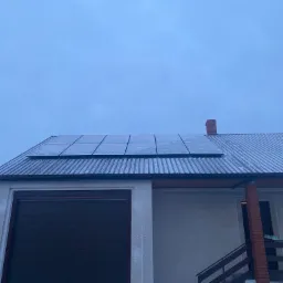 Panele słoneczne na metalowym dachu domu jednorodzinnego w pochmurny dzień, widoczne krople wody na panelach i dachu, z przodu garaż z brązową bramą.