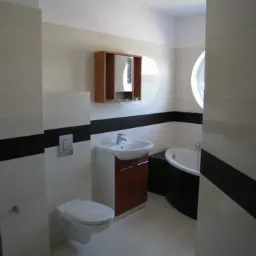 Nowoczesna łazienka z białymi i czarnymi płytkami, umywalką z szafką w kolorze drewna, wiszącą toaletą i okrągłym oknem nad wanną.