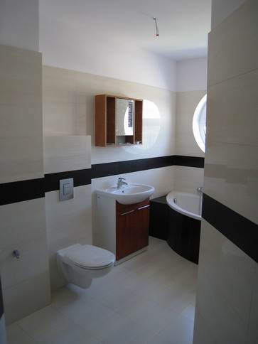 Nowoczesna łazienka z białymi i czarnymi płytkami, umywalką z szafką w kolorze drewna, wiszącą toaletą i okrągłym oknem nad wanną.