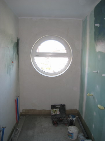 Remont łazienki w Pawłówku: widok na wnętrze w trakcie prac wykończeniowych, okrągłe okno, narzędzia i materiały budowlane na podłodze, częściowo położona gładź na ścianach.