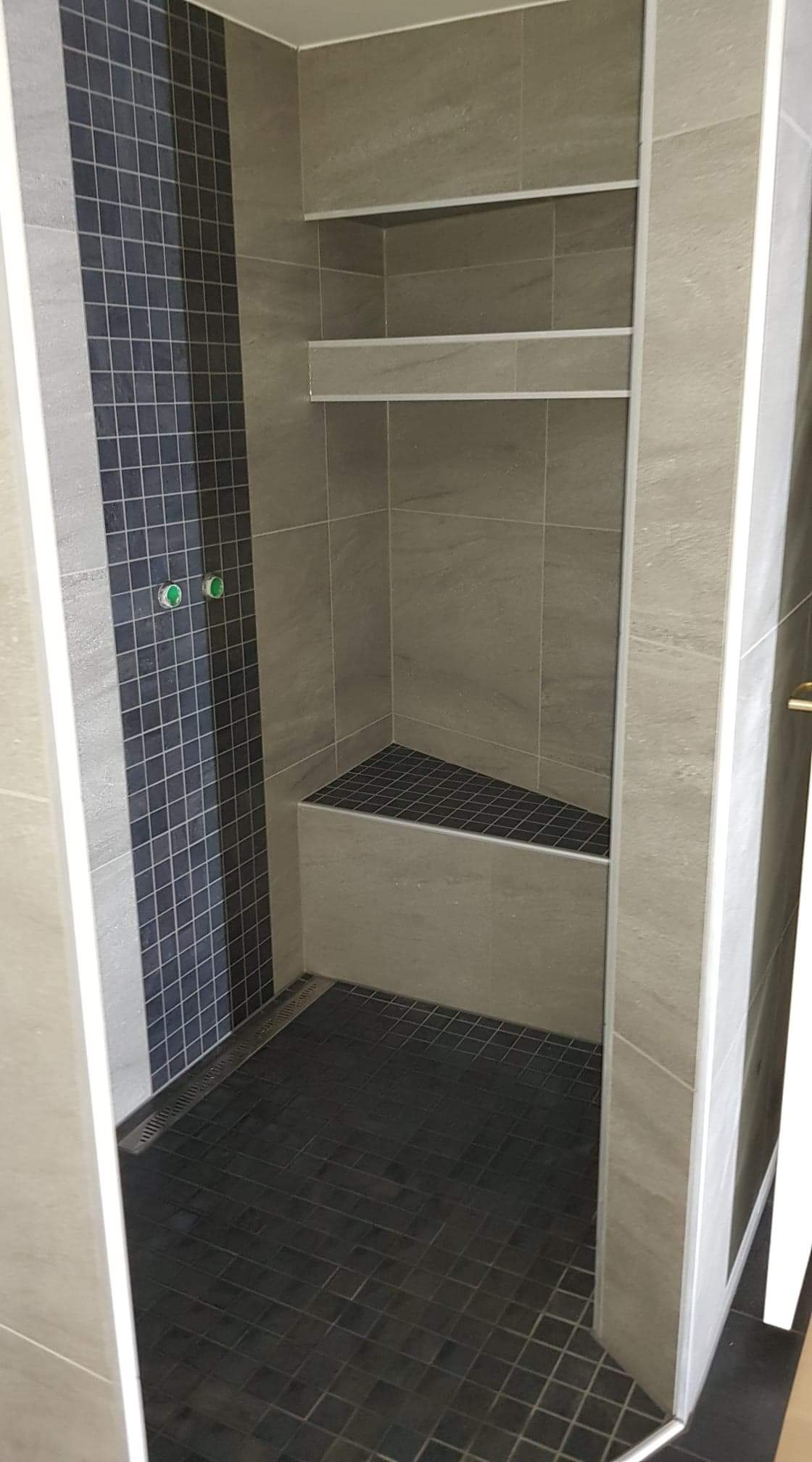 Nowoczesna kabina prysznicowa z szarymi płytkami imitującymi kamień, wnęką z półkami i siedziskiem wyłożonymi ciemną mozaiką, oraz odpływem liniowym w podłodze.