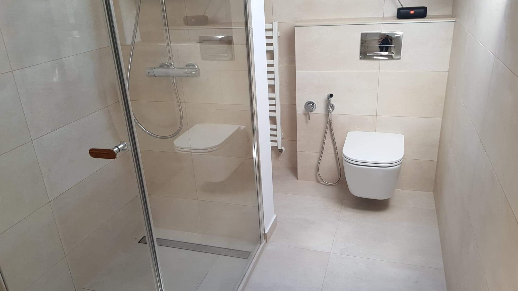 Nowoczesna łazienka z kabiną prysznicową ze szklanymi drzwiami z drewnianym uchwytem, podwieszaną toaletą z bidetką i grzejnikiem drabinkowym, wykończona beżowymi płytkami.