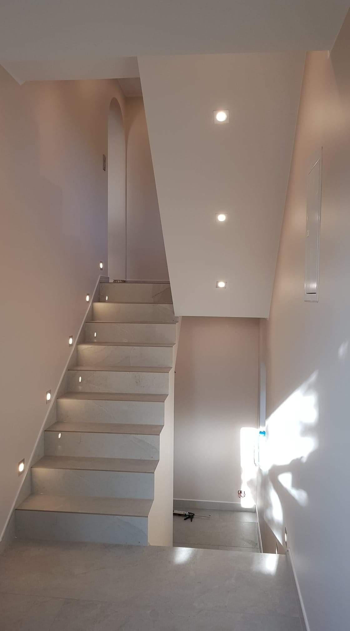 Wykończone schody z jasnego kamienia z podświetleniem LED w ścianie, widok z dołu na górę, świeżo po remoncie, z widocznym aplikatorem do silikonu na podłodze.