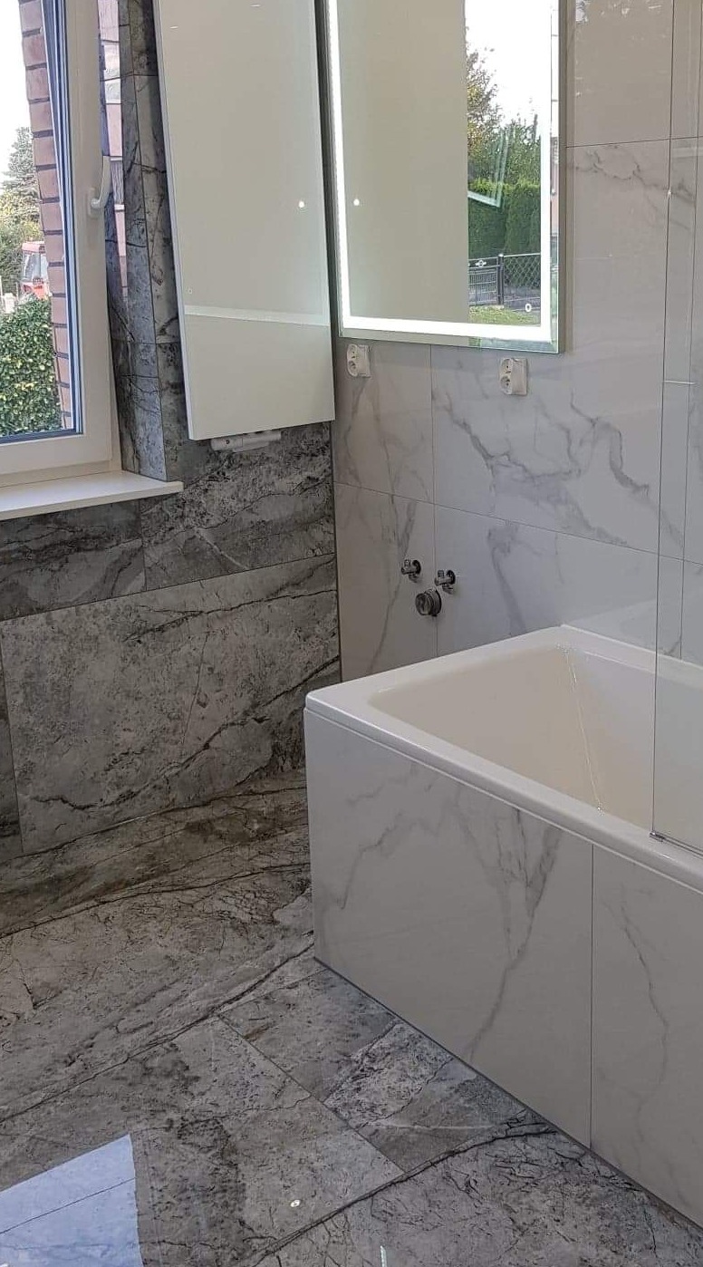 Nowoczesna łazienka z wanną obłożoną płytkami imitującymi marmur, podświetlane lustro i szare płytki podłogowe.