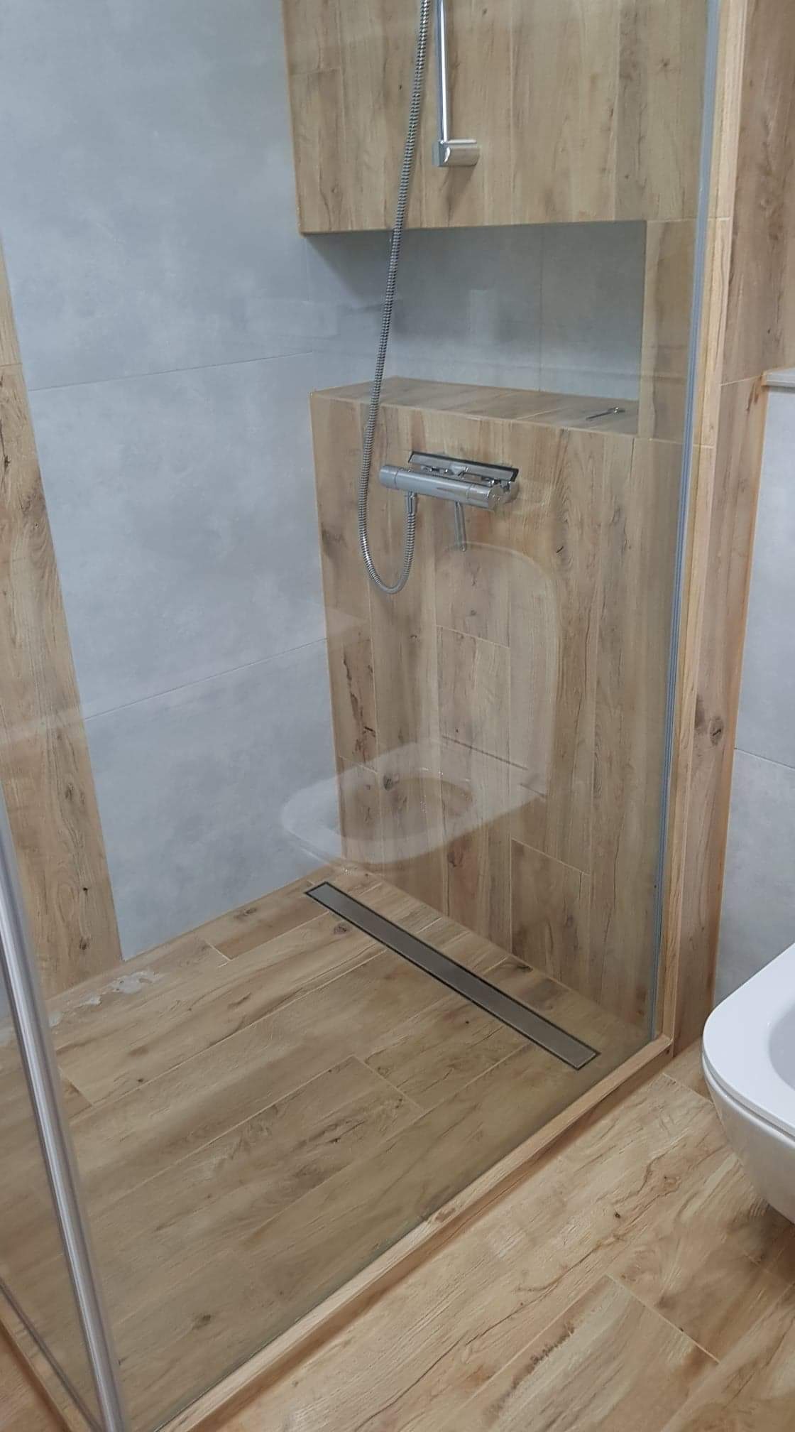 Nowoczesna kabina prysznicowa walk-in z odpływem liniowym, wykończona płytkami imitującymi drewno, szklana ścianka z chromowanymi profilami, widoczna część toalety.