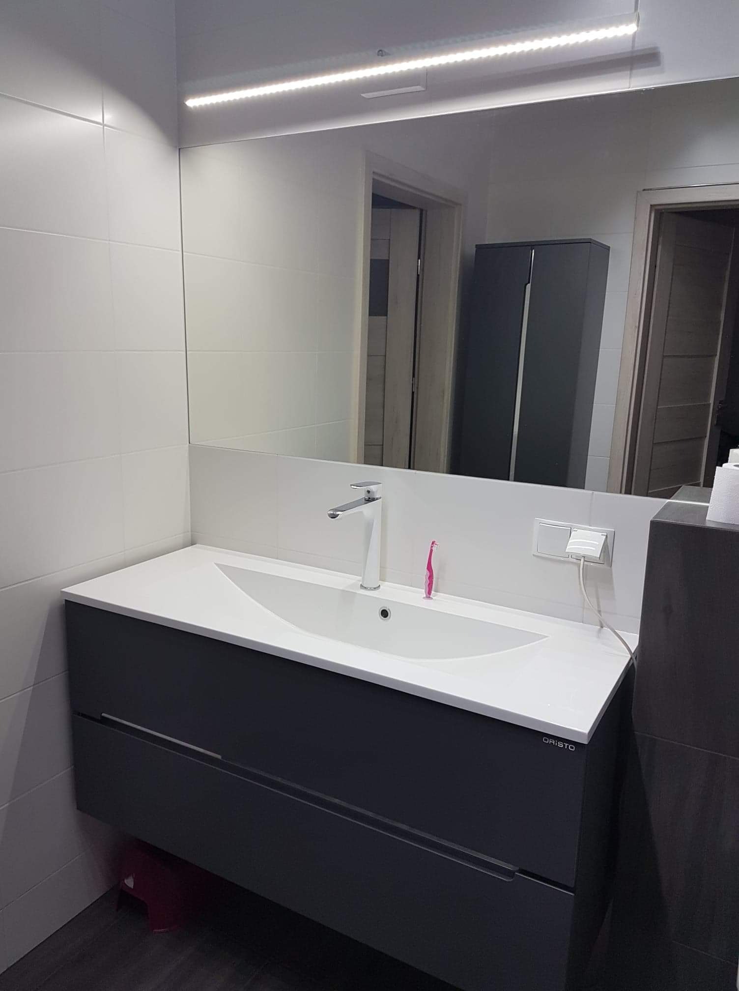 Nowoczesna łazienka z białą umywalką nablatową, szarą szafką pod umywalką z logo 'ORISTO', białymi płytkami na ścianach, podświetlanym lustrem i różową szczoteczką do zębów przy baterii.