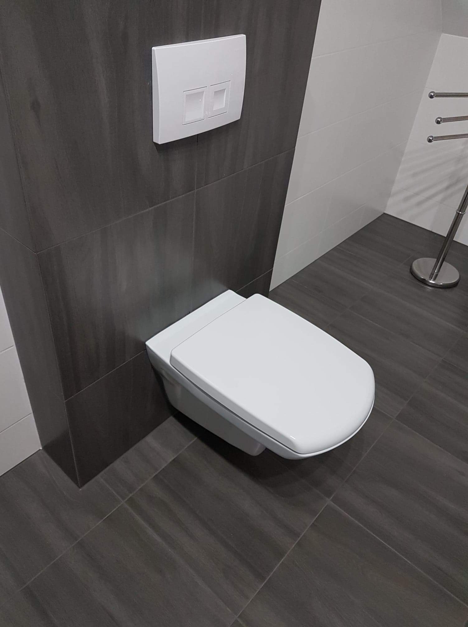 Podwieszana toaleta z przyciskiem spłukującym na ścianie wyłożonej ciemnymi, drewnopodobnymi płytkami, na podłodze identyczne płytki, fragment grzejnika drabinkowego w tle.