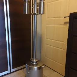 Metalowa kolumna rektyfikacyjna z zbiornikiem, wykonana ze stali nierdzewnej, stojąca na podłodze w pomieszczeniu mieszkalnym, widoczna na tle szafy i drzwi.