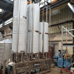 WeldingStal - Cztery wysokie, cylindryczne zbiorniki ze stali nierdzewnej, połączone rurami i umieszczone na metalowej ramie, w hali produkcyjnej z widocznym suwnicą i oknami dachowymi.