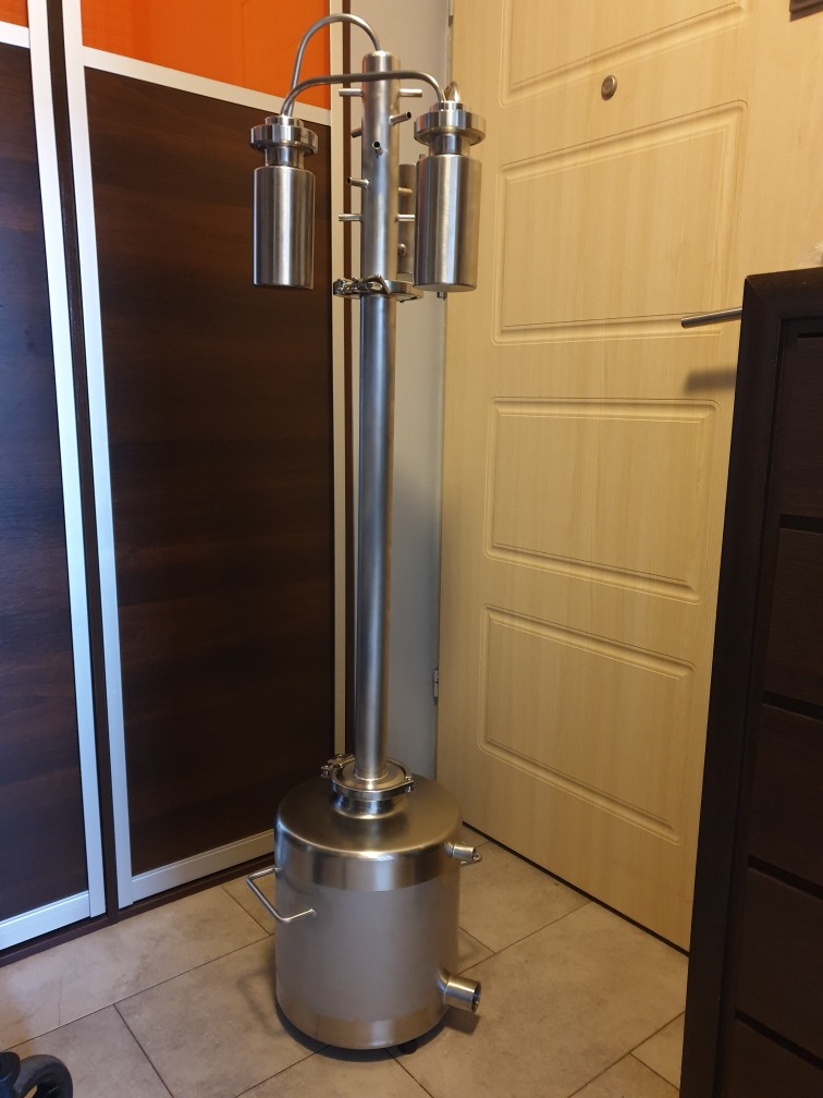 Metalowa kolumna rektyfikacyjna z zbiornikiem, wykonana ze stali nierdzewnej, stojąca na podłodze w pomieszczeniu mieszkalnym, widoczna na tle szafy i drzwi.