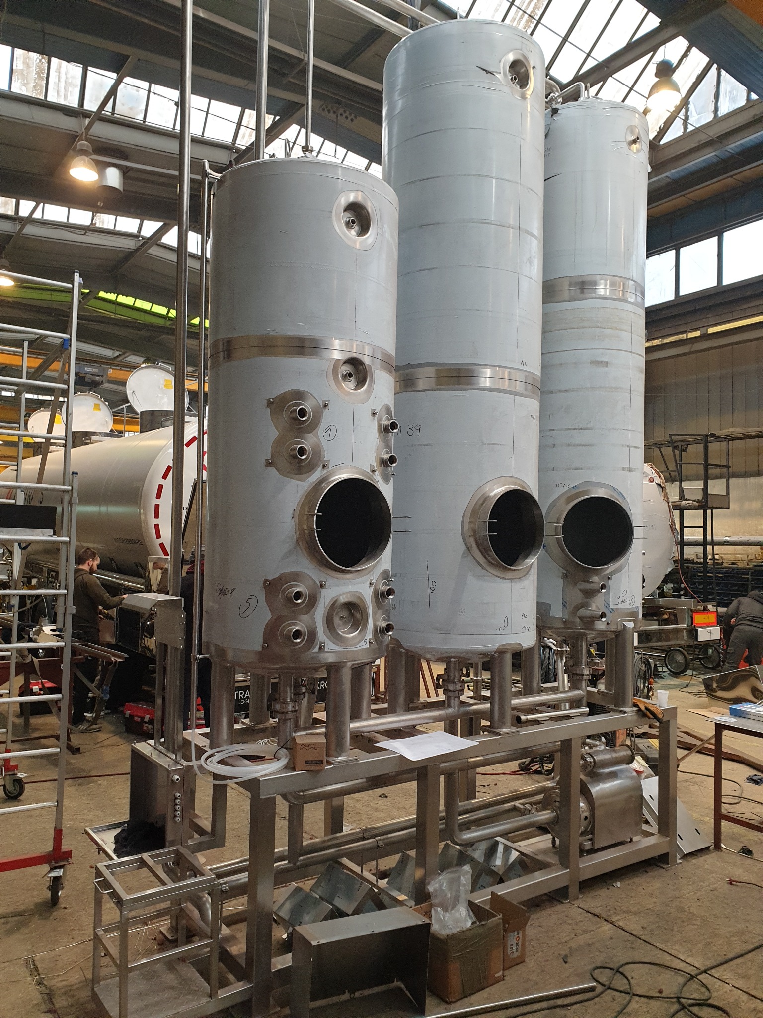 Trzy stalowe, cylindryczne zbiorniki przemysłowe w hali produkcyjnej, połączone systemem rur i ram stalowych, z widocznymi otworami inspekcyjnymi i portami przyłączeniowymi, w tle pracownik przy...