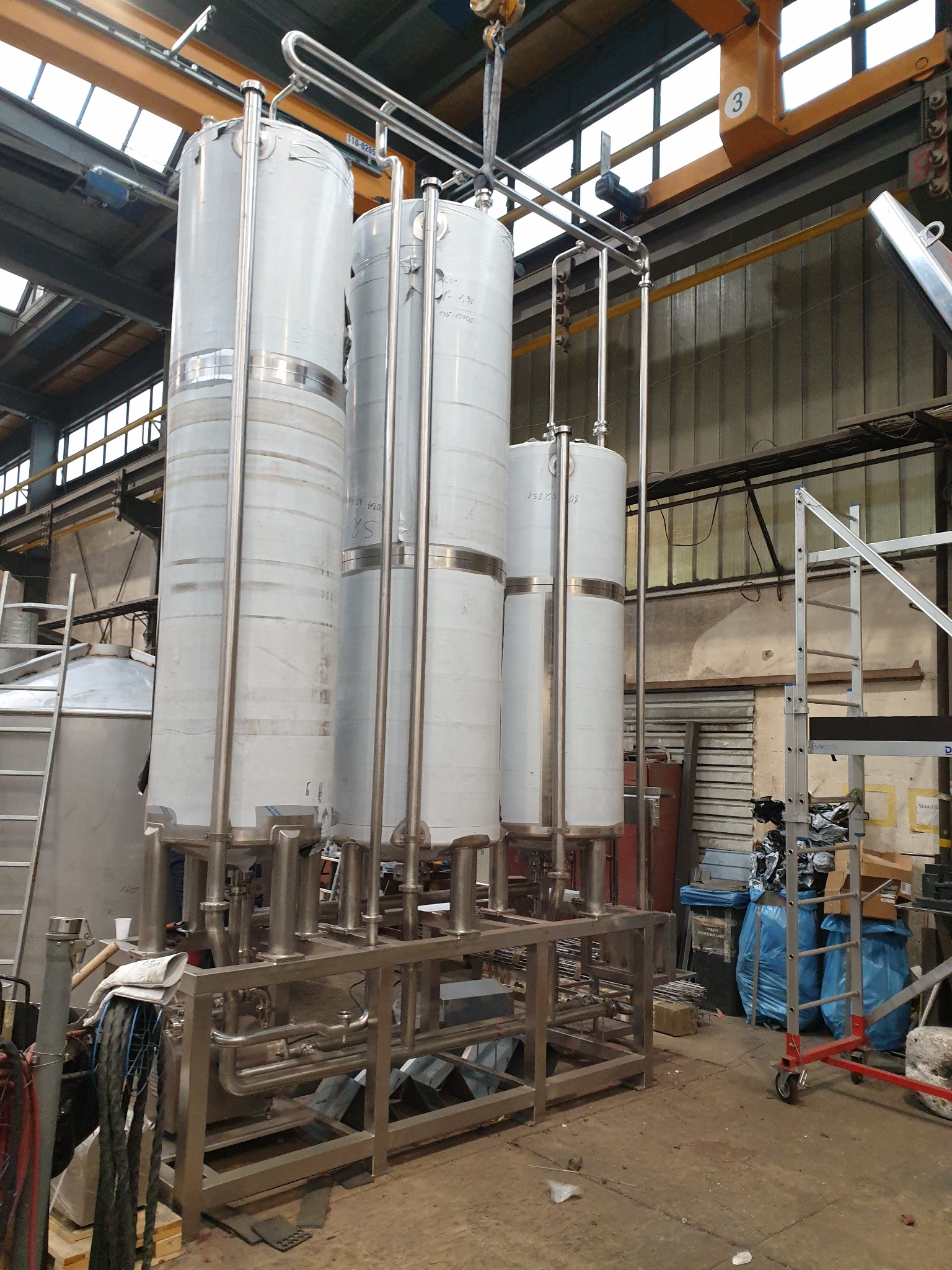Cztery wysokie, cylindryczne zbiorniki ze stali nierdzewnej, połączone rurami i umieszczone na metalowej ramie, w hali produkcyjnej z widocznym suwnicą i oknami dachowymi.