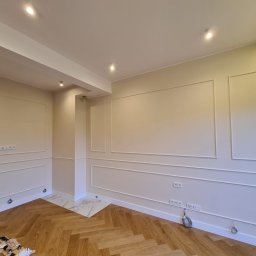 Wy-Remontowani - Wnętrze pokoju z parkietem ułożonym w jodełkę, kremowe ściany z dekoracyjnymi białymi listwami, białe gniazdka elektryczne i fragment marmurowej podłogi przy kolumnie.