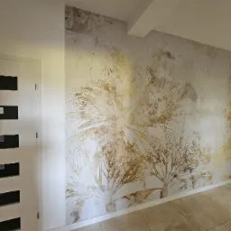 Ściana z dekoracyjną tapetą imitującą tropikalny motyw palm w odcieniach złota i beżu, widoczna obok białych drzwi z czarnymi poziomymi paskami, na podłodze płytki ceramiczne imitujące kamień.