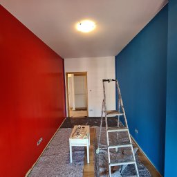 Wy-Remontowani - Pokój w trakcie odświeżania: ściana pomalowana na intensywny czerwony kolor kontrastuje z głębokim odcieniem niebieskiego, zabezpieczona podłoga, drabina oparta o niebieską ścianę, biały stolik...