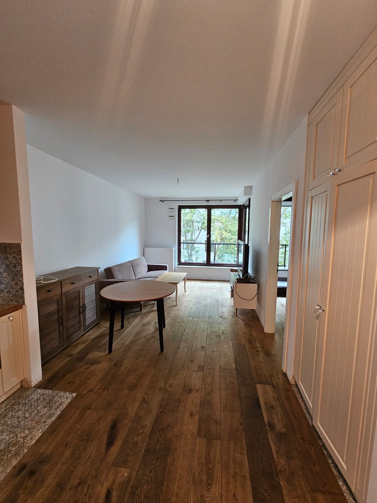 Jasne wnętrze mieszkania z drewnianą podłogą, okrągłym stołem i widokiem na okno z zielenią na zewnątrz. Szafa w zabudowie po prawej stronie, sofa w tle.