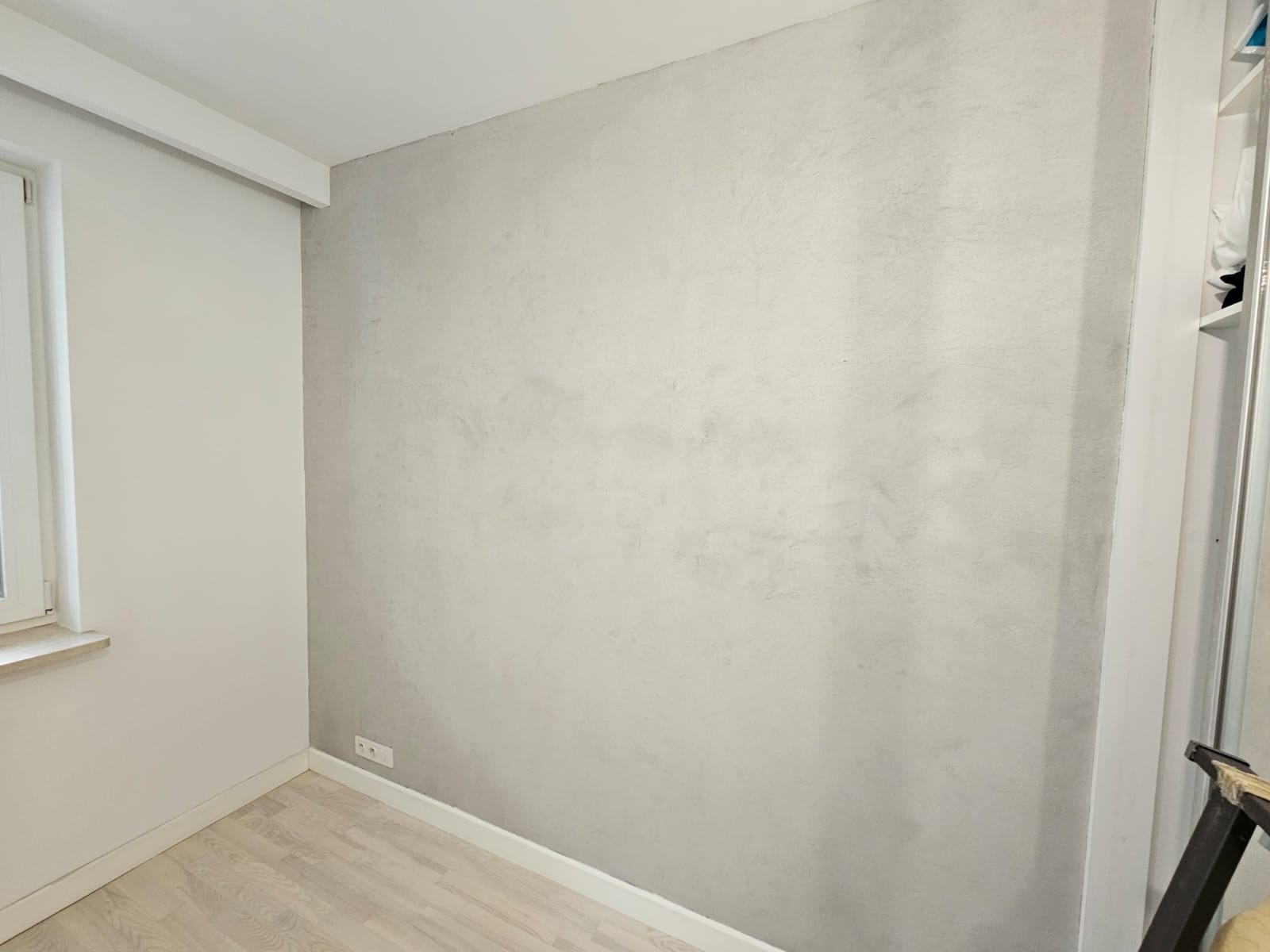 Ściana w pokoju wykończona dekoracyjną, szarą gładzią z efektem chmur, widoczne gniazdko elektryczne przy podłodze i fragment drewnianej podłogi.