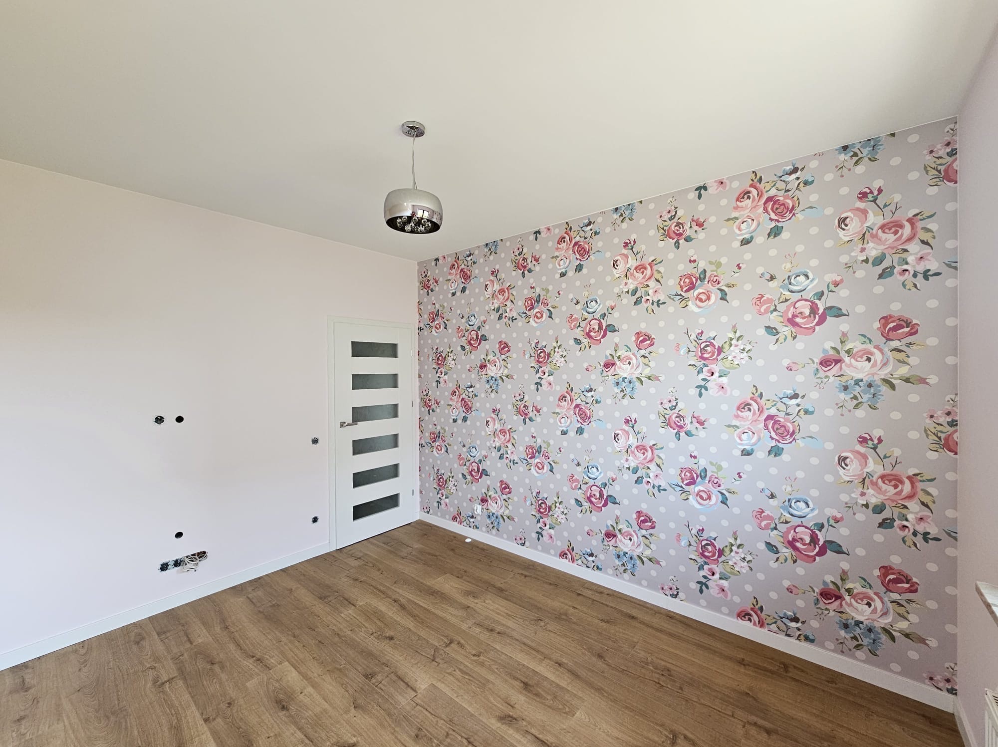 Pokój po odświeżeniu: jedna ściana z tapetą w kwiaty i kropki, pozostałe ściany w kolorze pudrowego różu, podłoga wyłożona panelami, widoczne gniazdka elektryczne bez osprzętu.