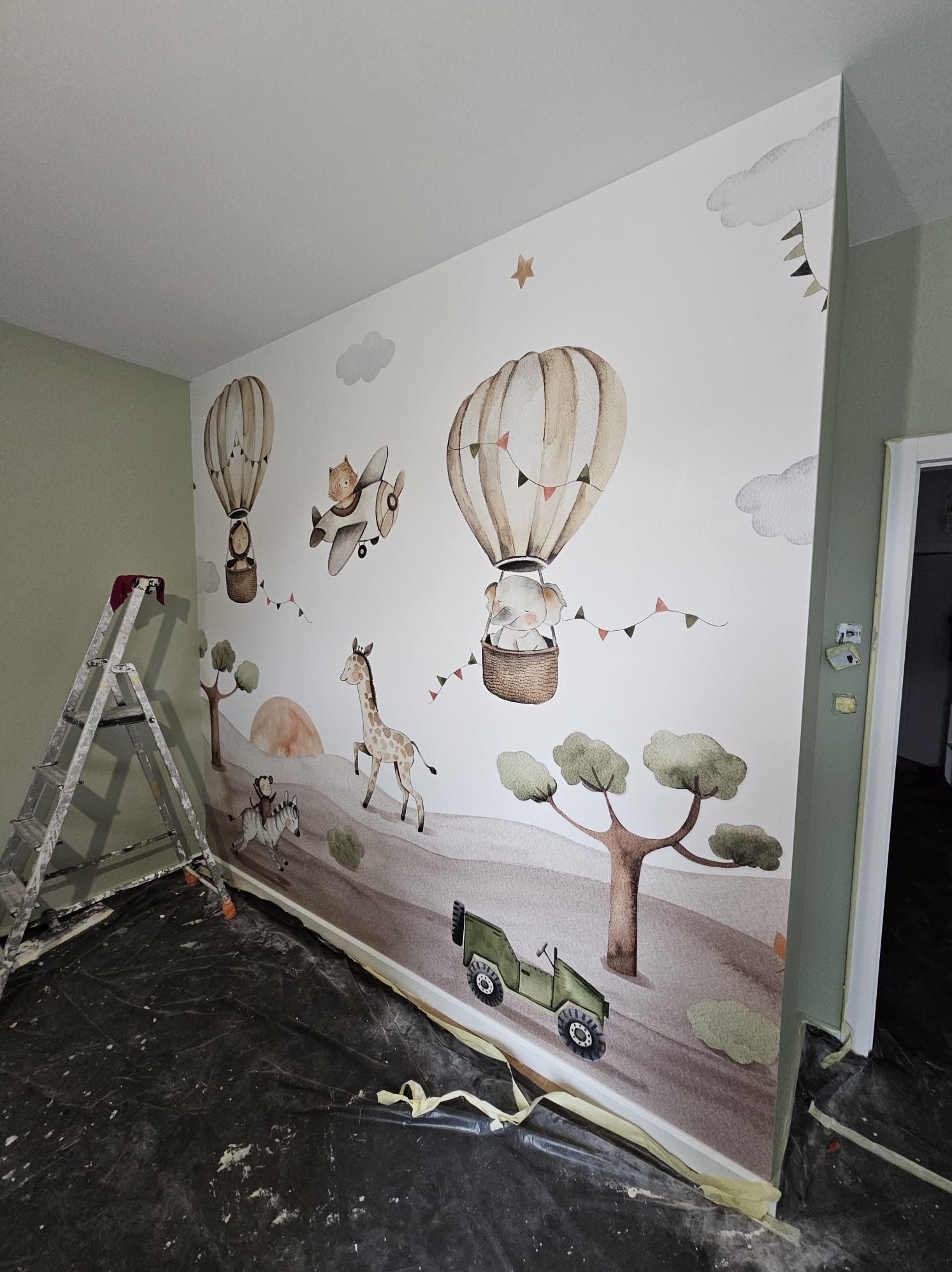Ściana pokoju dziecięcego z motywem safari: balony, żyrafa, zebra i jeep, częściowo zabezpieczona folią malarską, z widoczną aluminiową drabiną malarską po lewej stronie.
