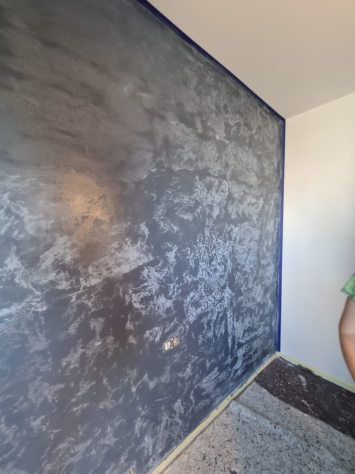 Ściana pokryta dekoracyjną, srebrzysto-szarą farbą z efektem ombre, zabezpieczona niebieską taśmą malarską przy suficie i narożnikach, widoczne gniazdko elektryczne i częściowo zakryta podłoga.