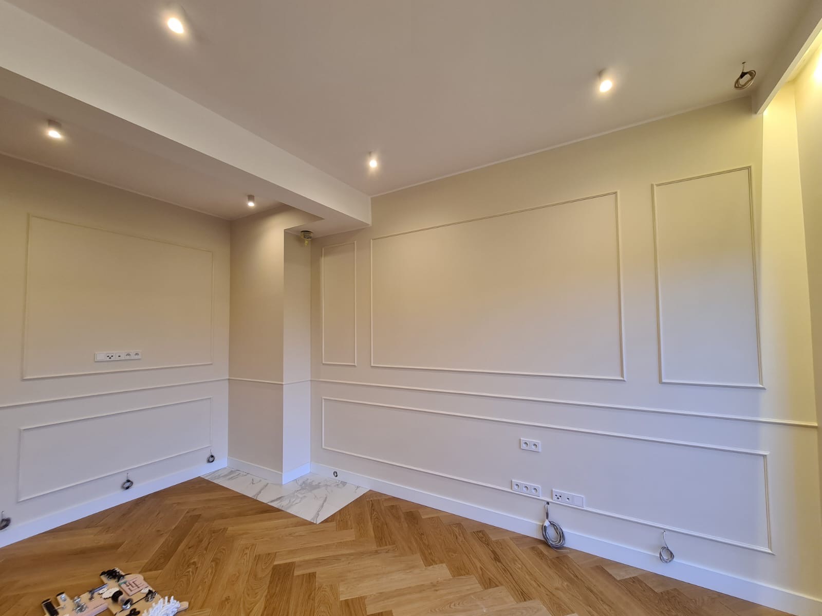 Wnętrze pokoju z parkietem ułożonym w jodełkę, kremowe ściany z dekoracyjnymi białymi listwami, białe gniazdka elektryczne i fragment marmurowej podłogi przy kolumnie.