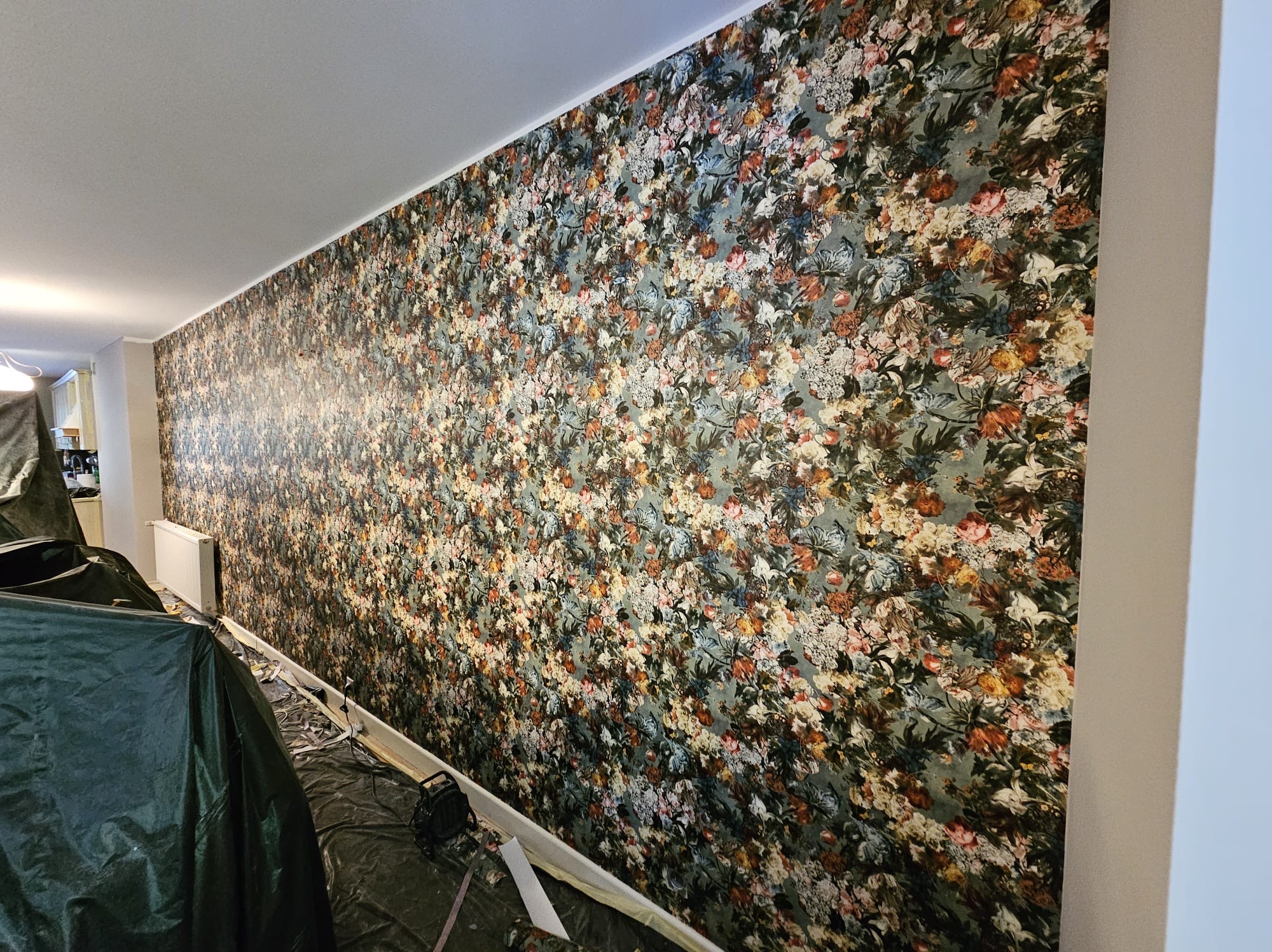 Ściana po położeniu wzorzystej tapety w kwiaty, zabezpieczona folią malarską, widoczny grzejnik i listwa przypodłogowa.