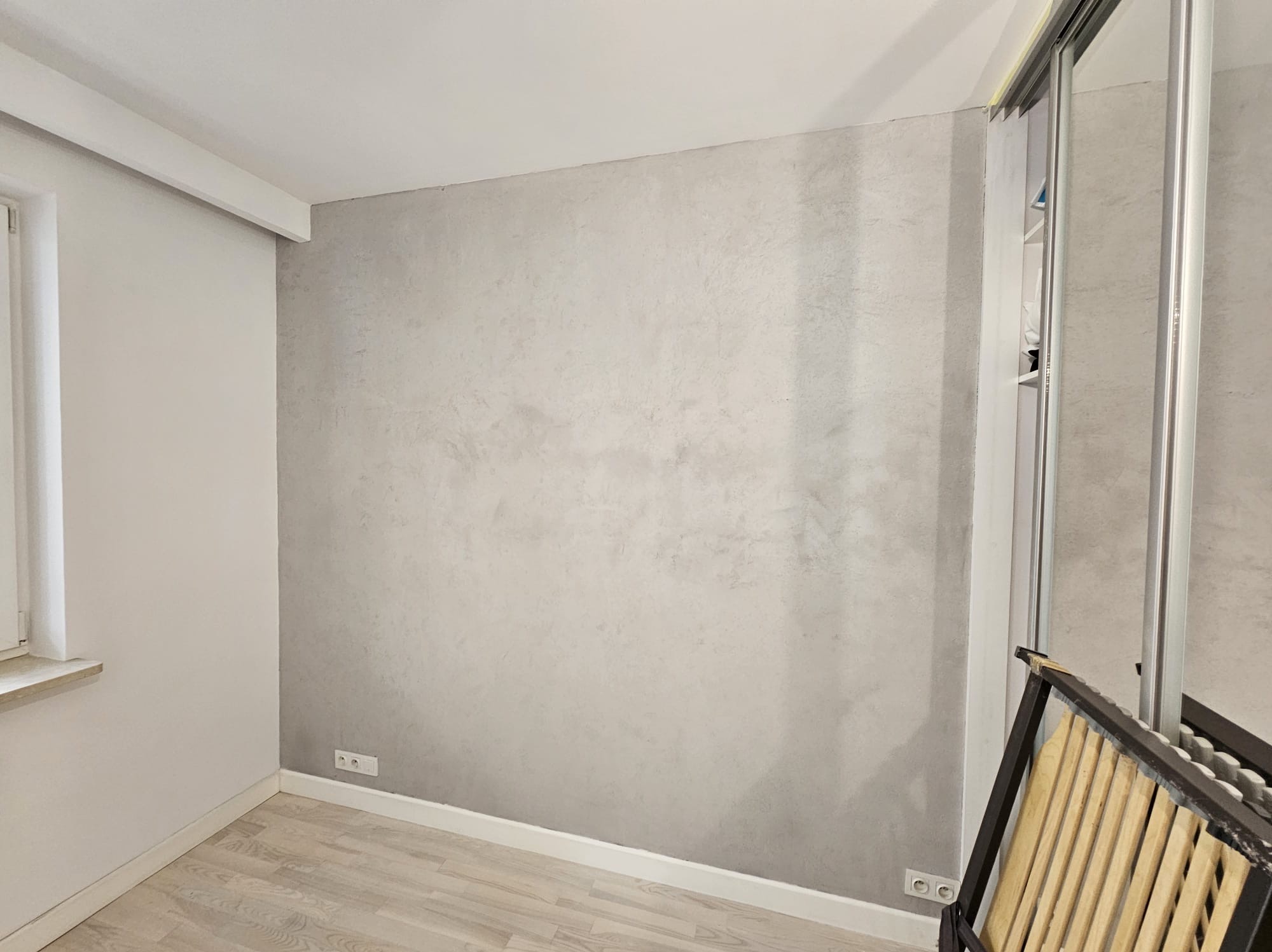 Ściana w pokoju po nałożeniu gładzi dekoracyjnej w odcieniach szarości, widoczne gniazdka elektryczne i fragment rozłożonego stelaża łóżka przy ścianie.