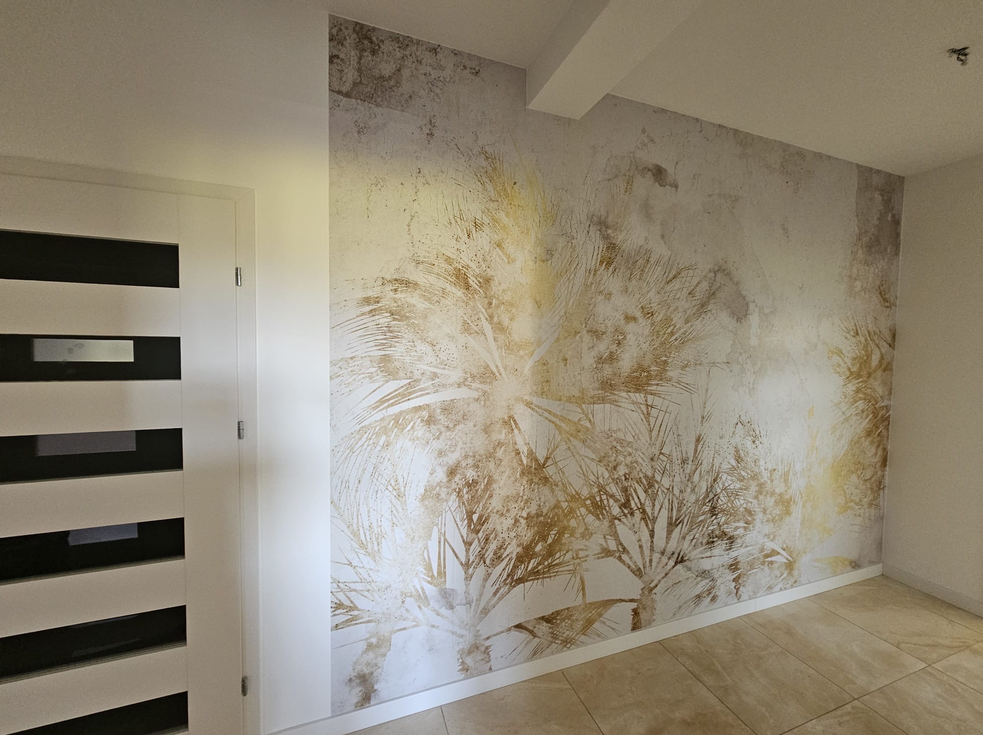 Ściana z dekoracyjną tapetą imitującą tropikalny motyw palm w odcieniach złota i beżu, widoczna obok białych drzwi z czarnymi poziomymi paskami, na podłodze płytki ceramiczne imitujące kamień.