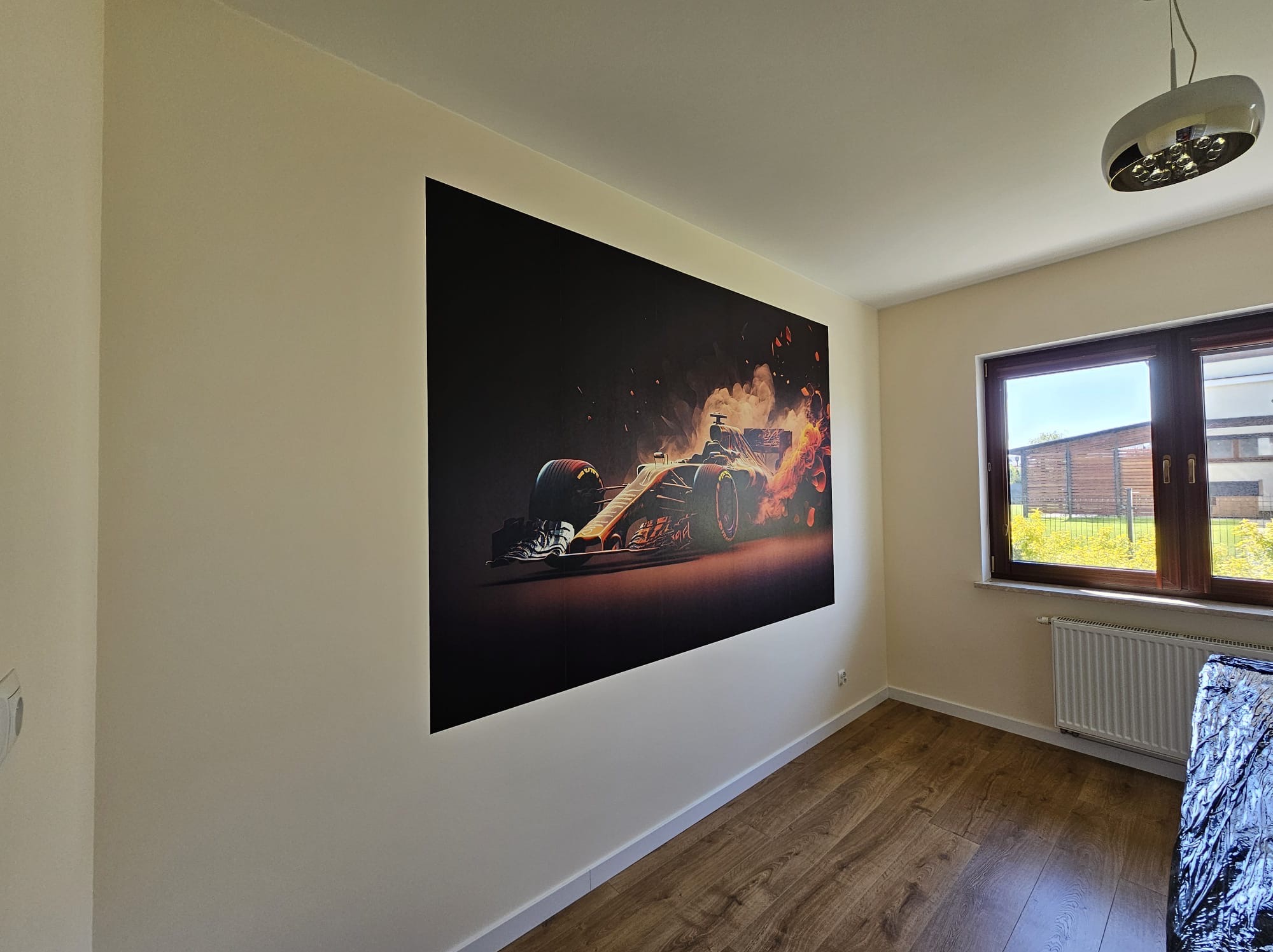 Duży plakat bolidu Formuły 1 na ścianie w pokoju z panelami podłogowymi, widok na okno z brązowymi ramami, jasne ściany i sufit, nowoczesna lampa na suficie.