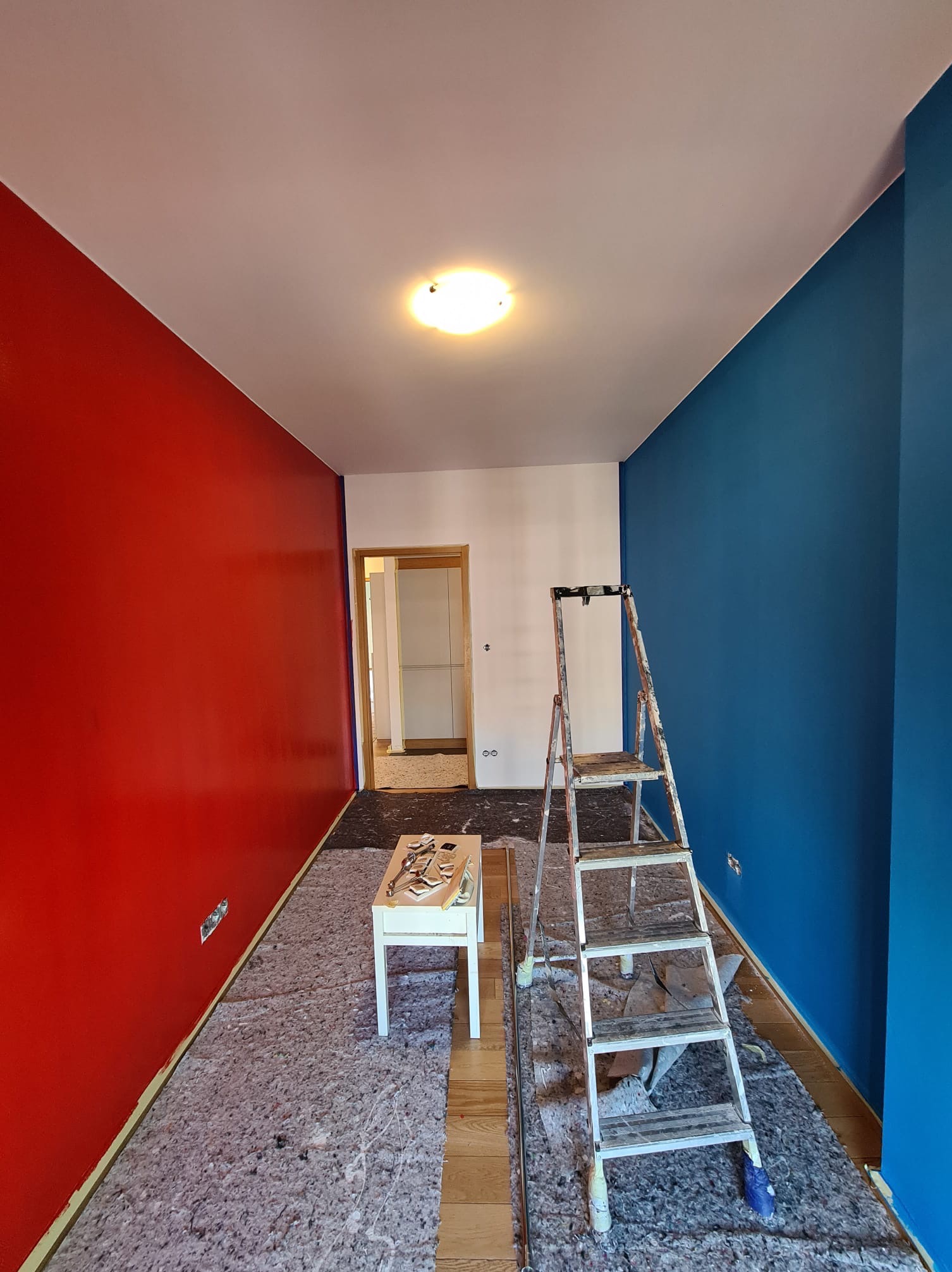 Pokój w trakcie odświeżania: ściana pomalowana na intensywny czerwony kolor kontrastuje z głębokim odcieniem niebieskiego, zabezpieczona podłoga, drabina oparta o niebieską ścianę, biały stolik...