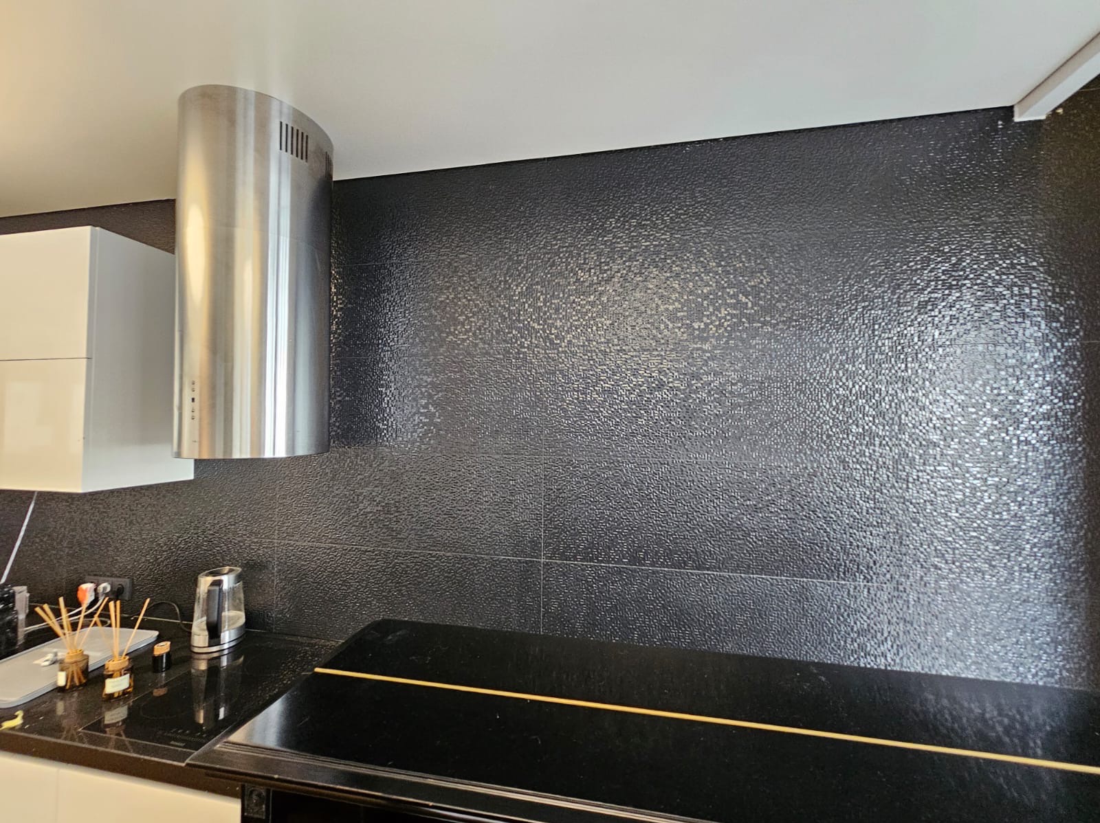 Część kuchni z czarnymi, teksturowanymi płytkami na ścianie, srebrnym okapem cylindrycznym i czarnym blatem z płytą grzewczą oraz białymi szafkami.