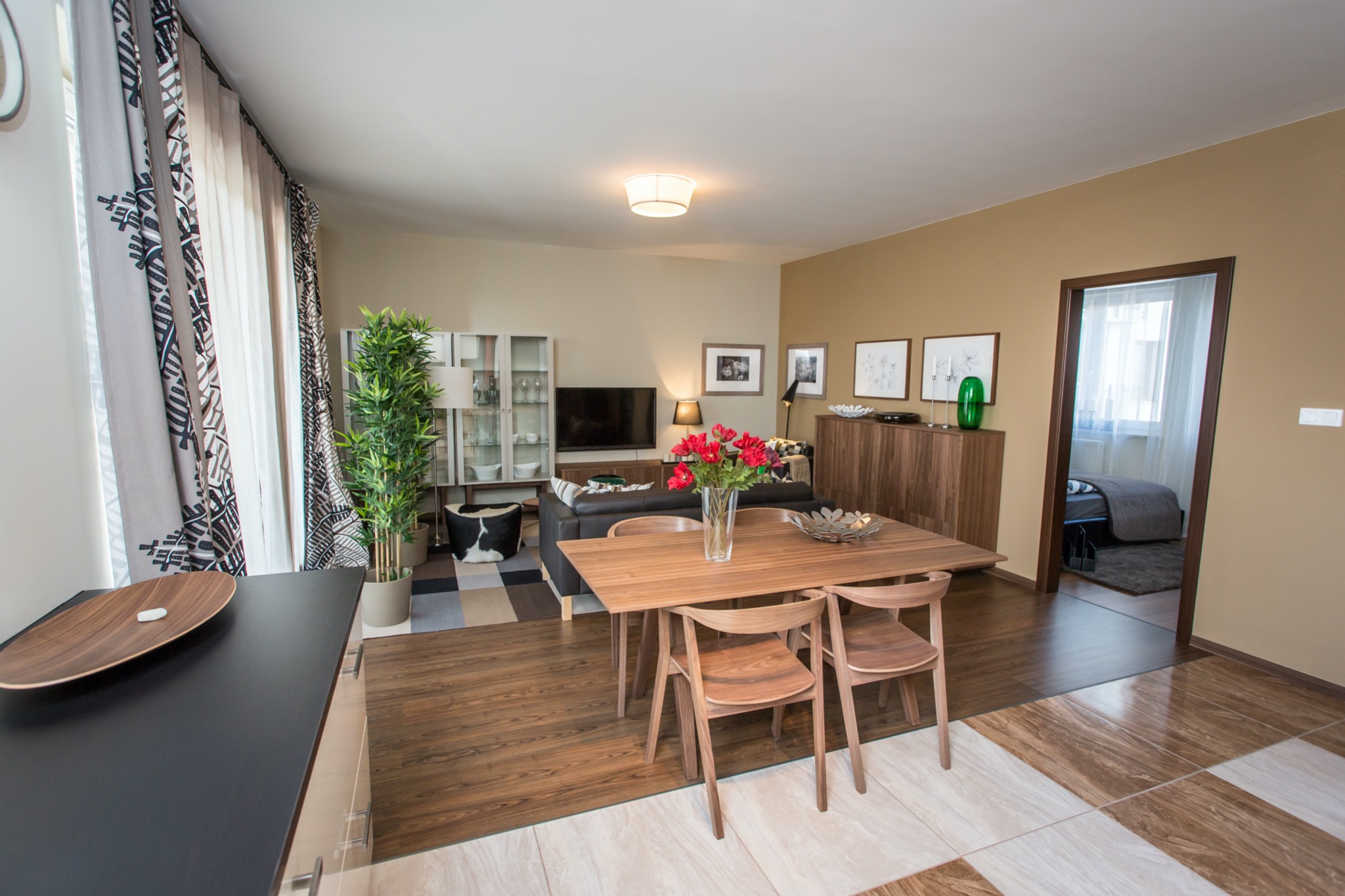 Jasne wnętrze mieszkania z drewnianym stołem i krzesłami, bukietem czerwonych róż na stole, widokiem na salon i sypialnię w tle, oraz wzorzystymi zasłonami po lewej stronie