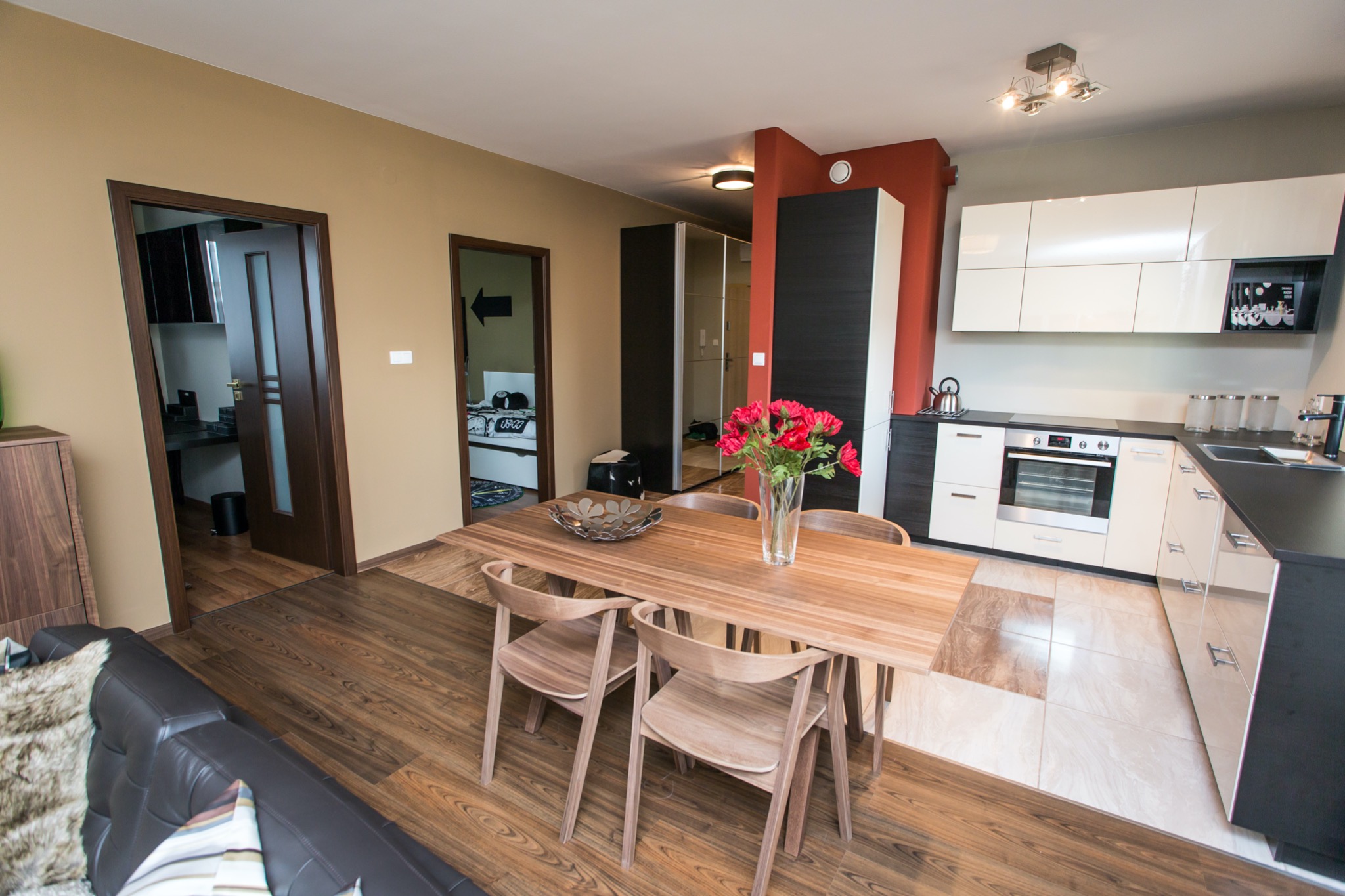 Jasne drewniane krzesła i stół w salonie z aneksem kuchennym, czerwone róże w wazonie, widok na sypialnię z łóżkiem i strzałką na ścianie.