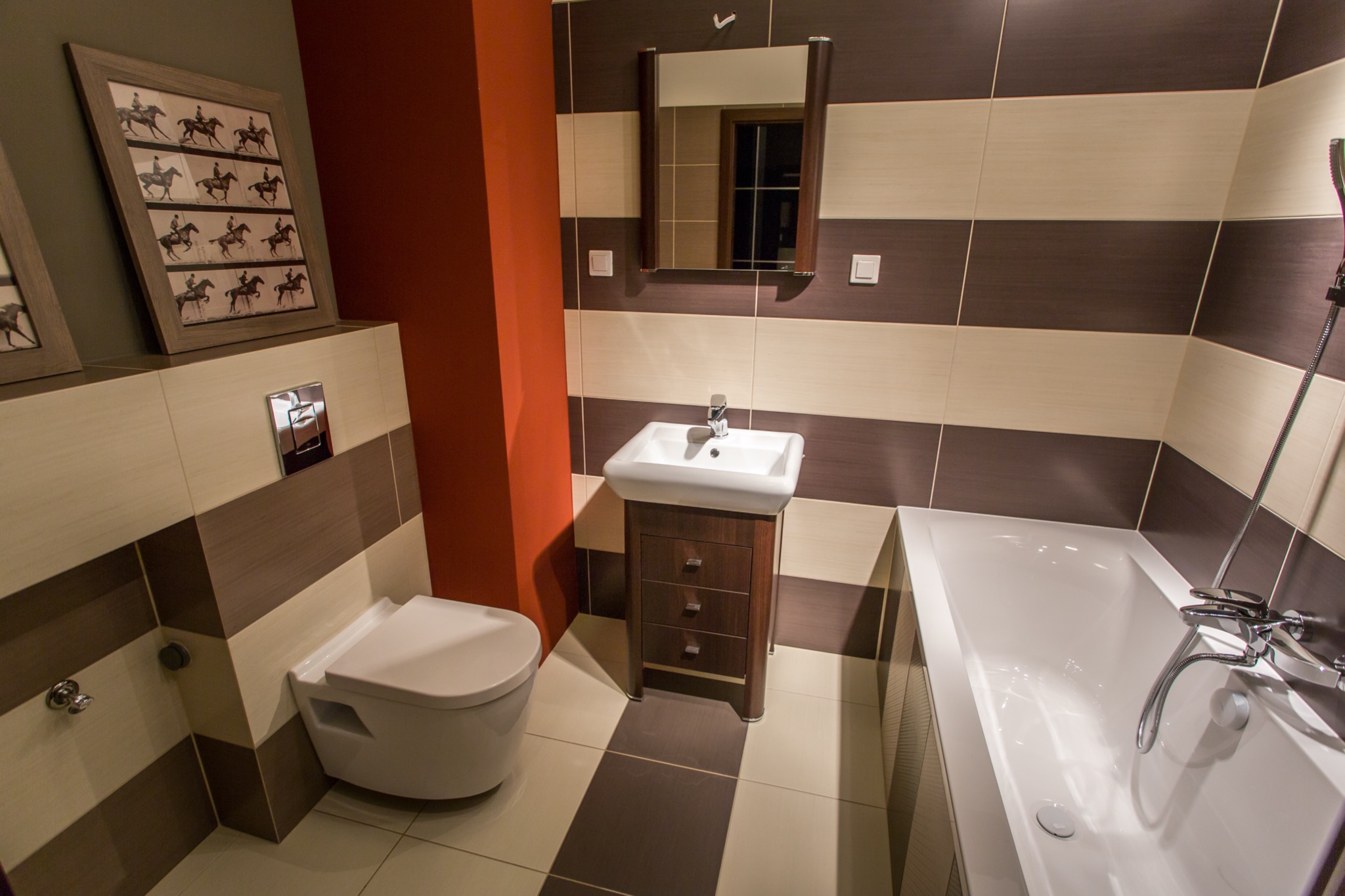 Nowoczesna łazienka z poziomymi pasami brązowo-kremowych płytek, wisząca toaleta, szafka pod umywalkę z trzema szufladami, lustro w drewnianej ramie i wanna, całość oświetlona ciepłym światłem.