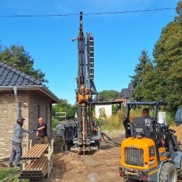 Geothermal Solutions P.S.A. - Wiercenie otworów pod gruntową pompę ciepła przy nowo wybudowanym domu z cegły, widoczna wiertnica, rury do pompy oraz mini ładowarka.