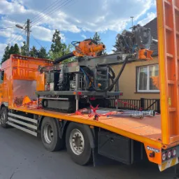 Pomarańczowy samochód ciężarowy z platformą transportową w Rossoszycy przewożący wiertnicę geotermalną na gąsienicach, zabezpieczoną czerwonymi pasami transportowymi.