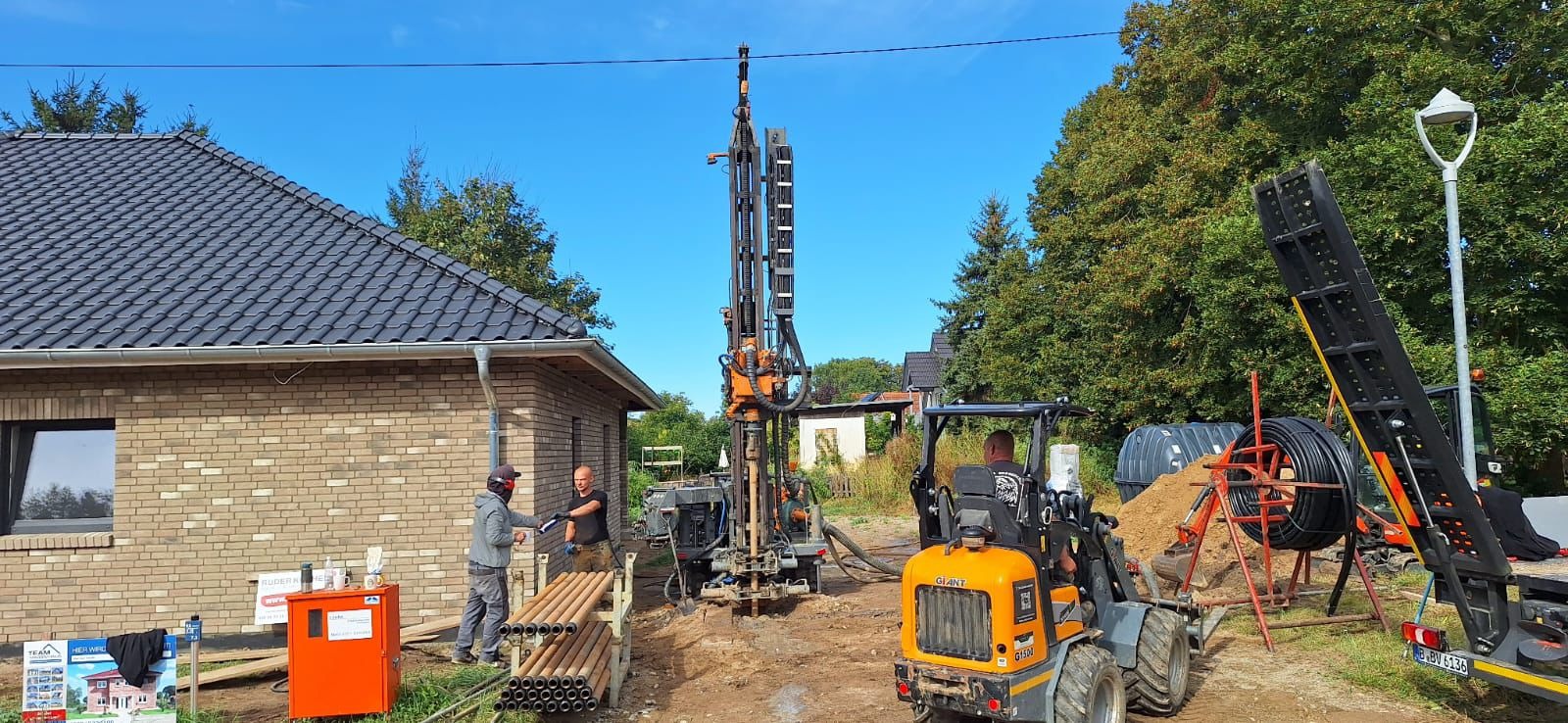 Wiercenie otworów pod gruntową pompę ciepła przy nowo wybudowanym domu z cegły, widoczna wiertnica, rury do pompy oraz mini ładowarka.