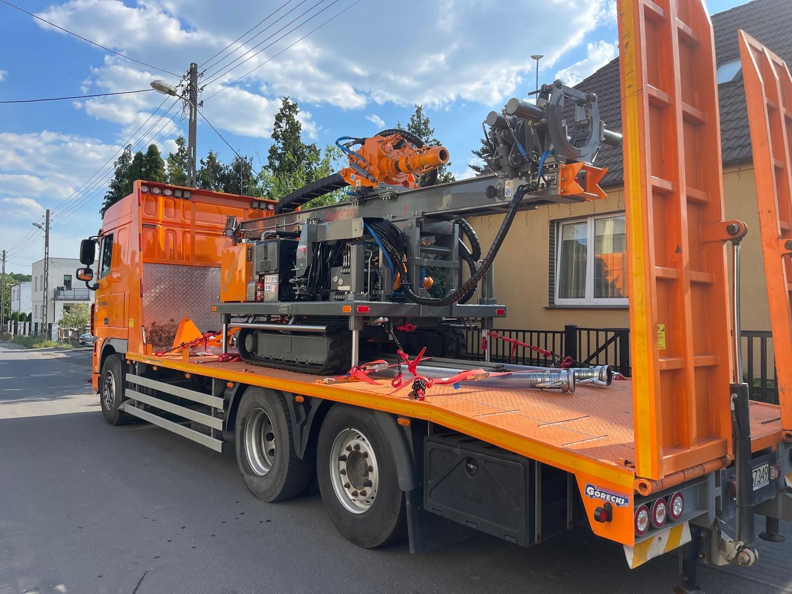 Pomarańczowy samochód ciężarowy z platformą transportową w Rossoszycy przewożący wiertnicę geotermalną na gąsienicach, zabezpieczoną czerwonymi pasami transportowymi.