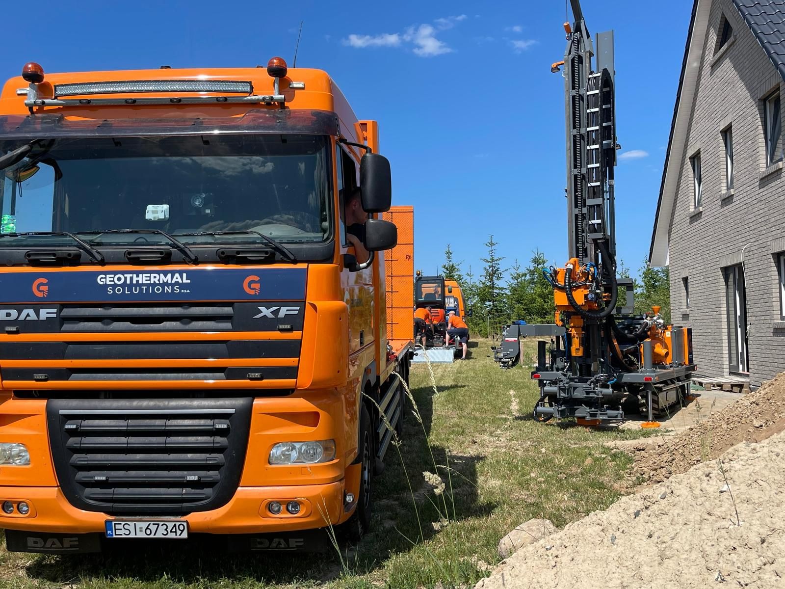 Pomarańczowa ciężarówka z logo firmy w trakcie wiercenia odwiertu pionowego pod instalację pompy ciepła obok domu z szarej cegły.