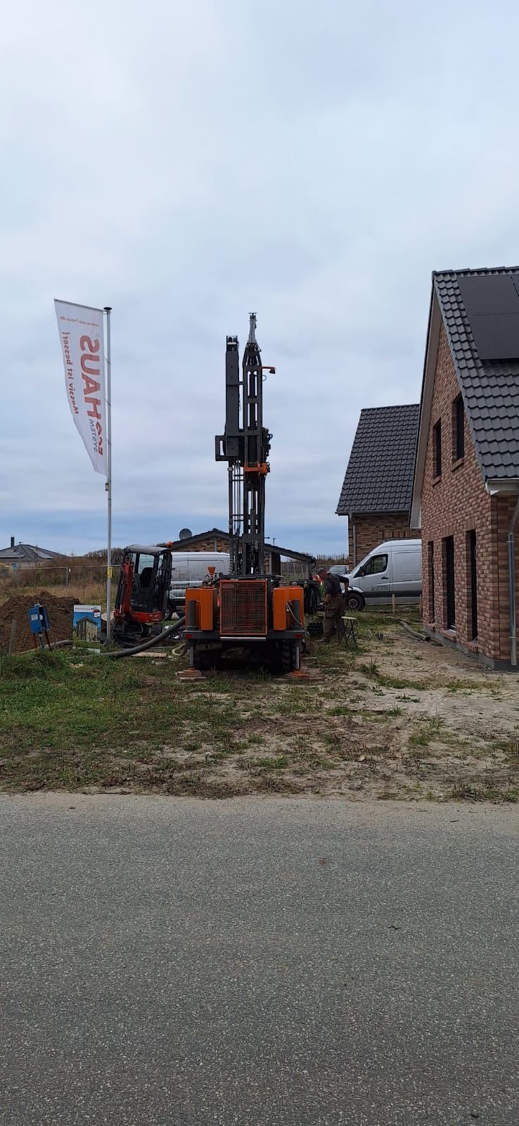 Wiertnica geotermalna w trakcie pracy na posesji z nowym domem z cegły i panelami słonecznymi na dachu, widoczny baner reklamowy firmy.