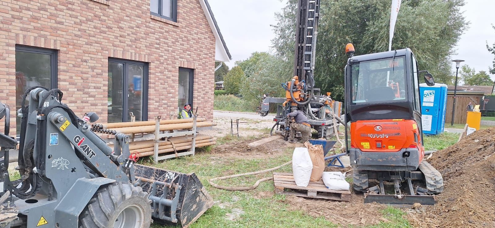 Wiertnica i minikoparka Kubota na placu budowy obok nowego, ceglanego domu podczas wiercenia otworów pod instalację gruntowej pompy ciepła. Widoczni pracownicy w trakcie pracy.