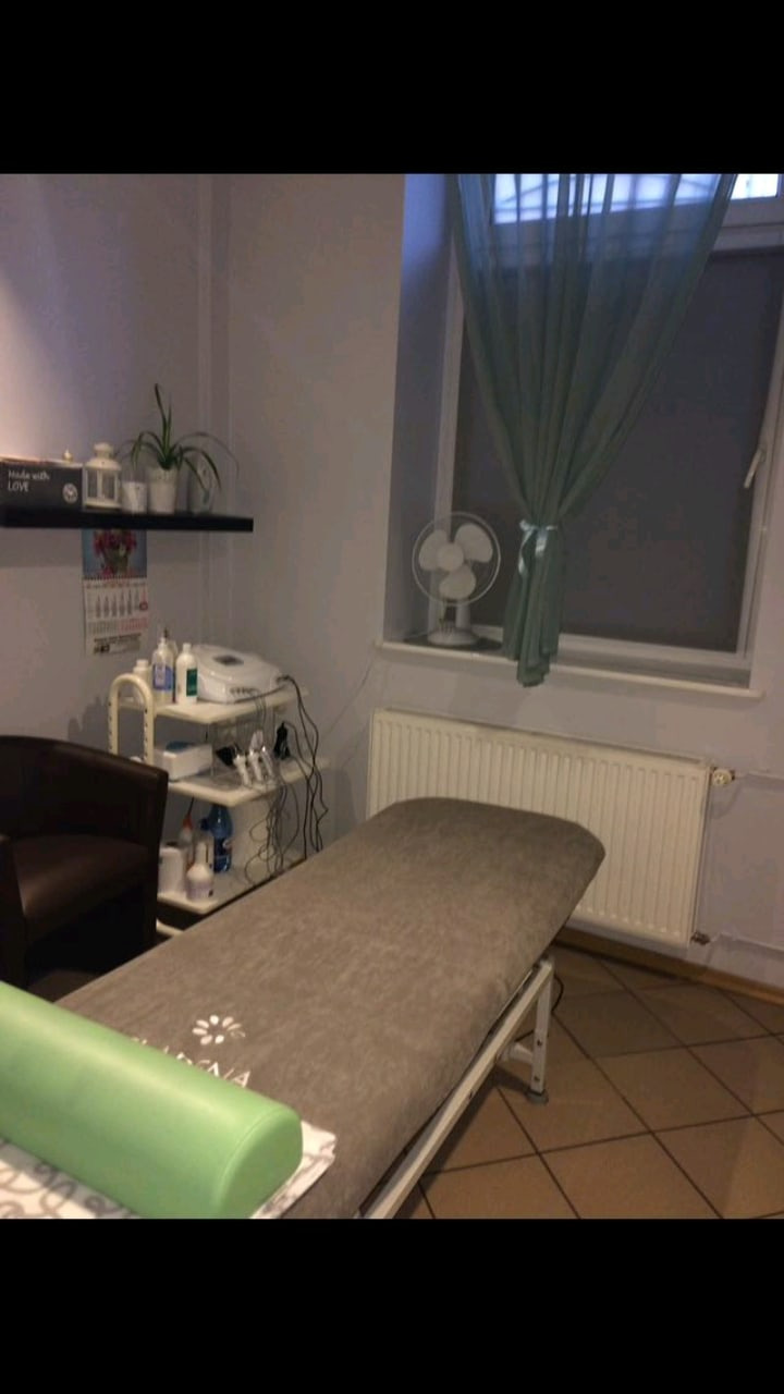 Gabinet z beżowym leżankiem do masażu, zielonym wałkiem, białym wózkiem z kosmetykami i wentylatorem przy oknie.