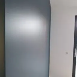 Pomieszczenie po malowaniu: gładka, szara ściana z delikatnym refleksem światła, kontrastująca z białą ścianą i fragmentem drewnianej podłogi. Widoczny fragment czarnej ramy drzwi.