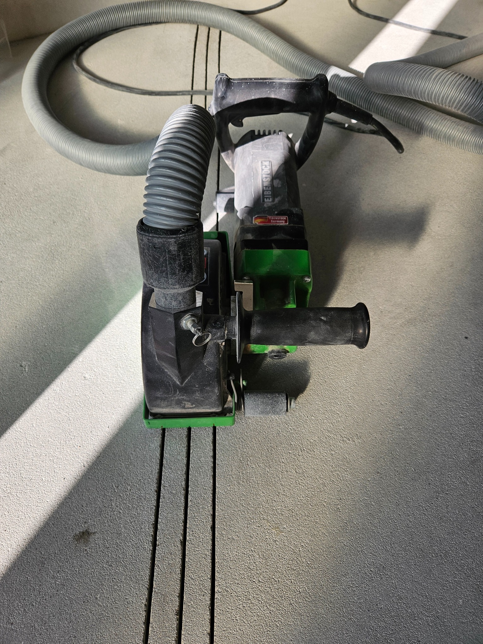 Profesjonalna bruzdownica Eibenstock z podłączonym odkurzaczem przemysłowym, przygotowana do wycinania rowków w posadzce, widoczne trzy wycięte bruzdy.