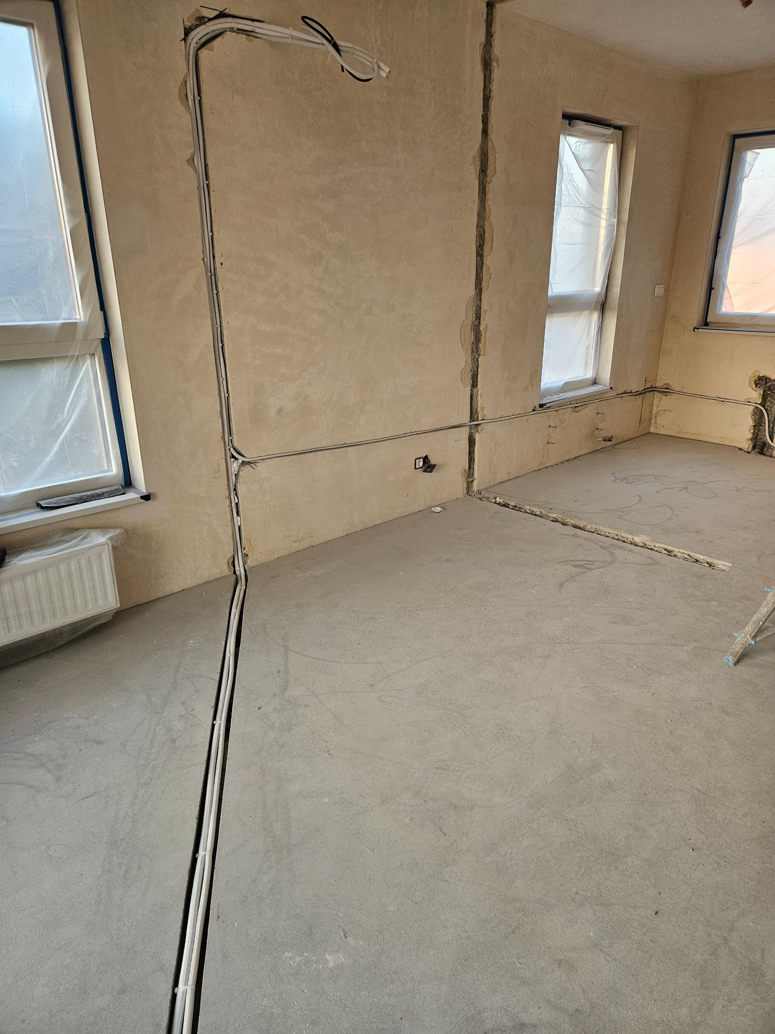 Wnętrze w stanie surowym z widocznymi bruzdami w ścianach i podłodze, w których poprowadzone są białe rurki instalacyjne. Okna zabezpieczone folią malarską.