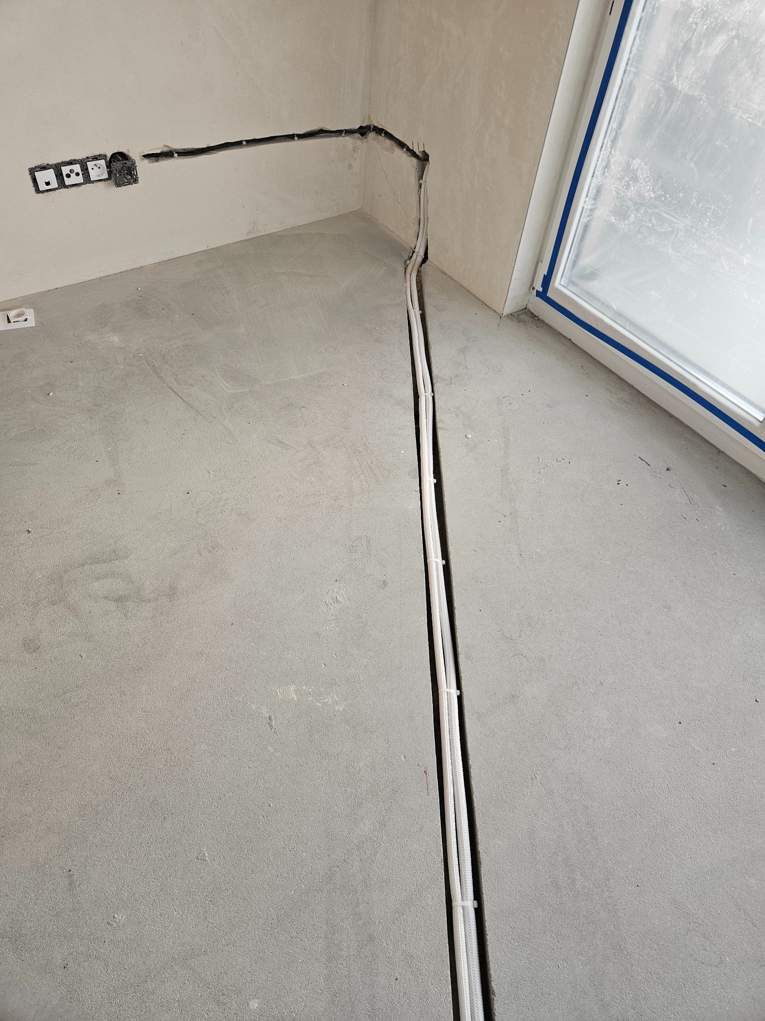 Wnętrze w trakcie remontu: kable elektryczne prowadzone w bruzdach w ścianie i podłodze, puszki elektryczne w ścianie, okno zabezpieczone taśmą malarską.