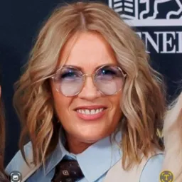 Portret uśmiechniętej kobiety w okularach, blond włosach i koszuli z krawatem, na tle ciemnego tła z logotypem. Kadr od ramion w górę, z widocznym fragmentem drugiej osoby.