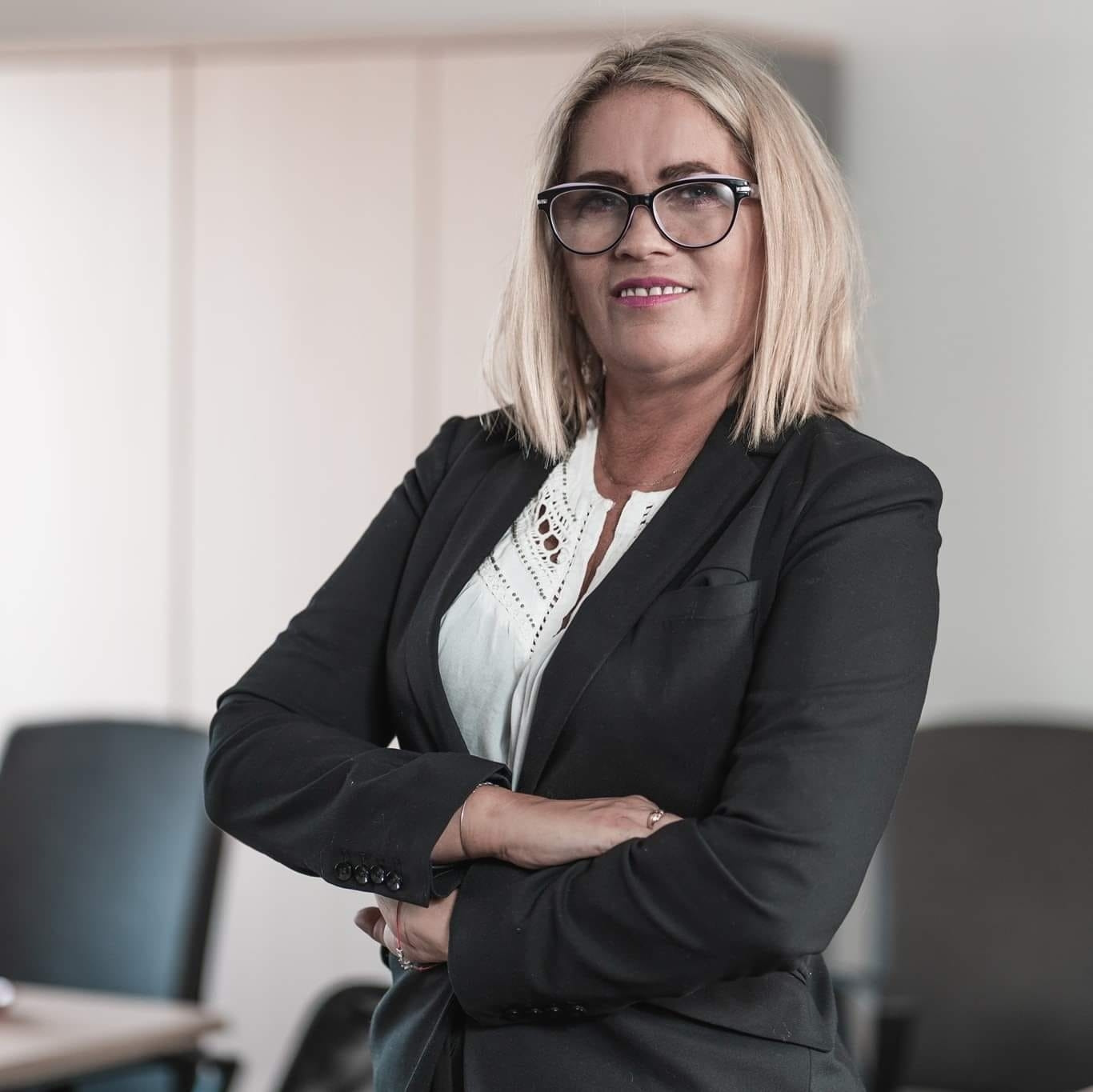 Kobieta w czarnej marynarce i okularach, z założonymi rękami, w jasnym biurze.